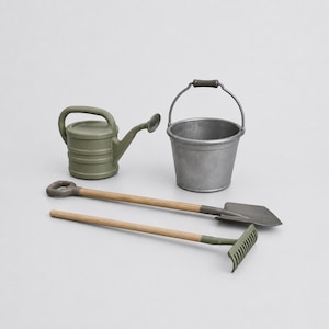 May include: A set of gardening tools including a matte olive green watering can, a metallic silver bucket with a dark handle, a shovel with a wooden handle and dark head, and a rake with a wooden handle and olive green head.