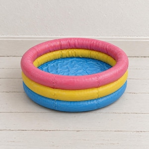 Peut inclure: Une piscine gonflable circulaire composée de trois anneaux horizontaux distincts aux couleurs vives. L'anneau supérieur est rose, l'anneau central est jaune et l'anneau inférieur est bleu. Le fond intérieur est une surface bleue texturée.