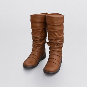 Puede incluir: Un par de botas de media caña en color marrón claro sólido. Estas botas presentan un diseño de caña fruncida y una serie de tiras horizontales que envuelven la zona del tobillo, terminadas con una suela plana y una puntera redondeada.