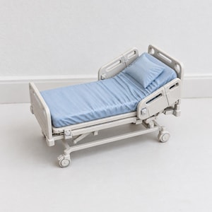May include: A miniature hospital bed featuring a light beige frame with adjustable side rails and a headboard. The bed is dressed with a light blue mattress and a matching rectangular pillow, supported by a wheeled base with four small casters.