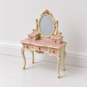 May include: A miniature vanity table featuring a cream-colored frame with pink accents, an oval mirror, and five small drawers. The piece is decorated with sculpted pink rose details and gold-toned knobs, standing on four curved legs.