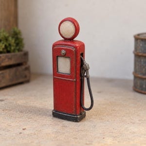 May include: A miniature red gas pump figurine featuring a circular top sign, a square display window, and a black hose with a nozzle attached to the side. The vintage-style pump has a weathered finish and sits on a rectangular black base.