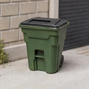 May include: A miniature green waste bin featuring a black hinged lid with a rectangular opening. The container has a molded rectangular body with vertical structural ribs and a single visible black wheel attached to the side of the base.