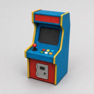 May include: A miniature arcade cabinet featuring a blue frame with yellow trim, a black screen, a red joystick, and six colourful buttons. The lower section is red with a silver coin door panel, and the top header area is a solid red rectangle.