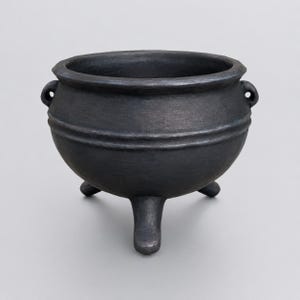 May include: A matte black cauldron with a rounded body, a wide circular opening, and three short, sturdy legs. The vessel features two small, integrated loop handles on opposite sides and a horizontal ridge detail encircling the upper section.