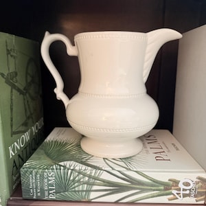 May include: A white ceramic pitcher with a curved handle, a wide spout, and a rounded body featuring a subtle beaded texture around the neck and base. The pitcher has a smooth, glossy finish and sits on a pedestal base, showcasing a classic design.