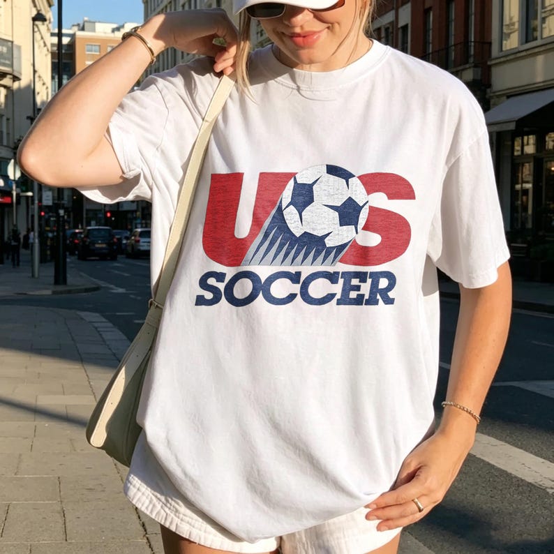 May include: A white short-sleeved t-shirt featuring a graphic print with the red letters US, a black and white soccer ball replacing the letter S, and the word SOCCER in blue capital letters underneath the graphic.