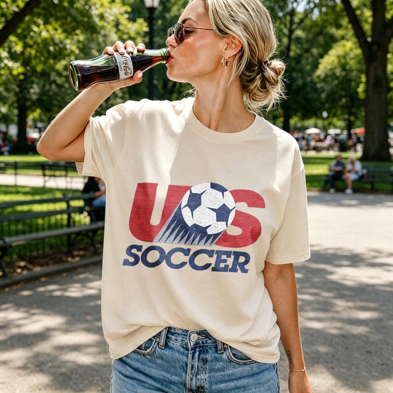 May include: A cream-colored short-sleeve t-shirt featuring a graphic print with the red text US, a soccer ball replacing the letter S, and the word SOCCER in blue text below. The shirt is paired with blue denim jeans and a glass bottle of soda.