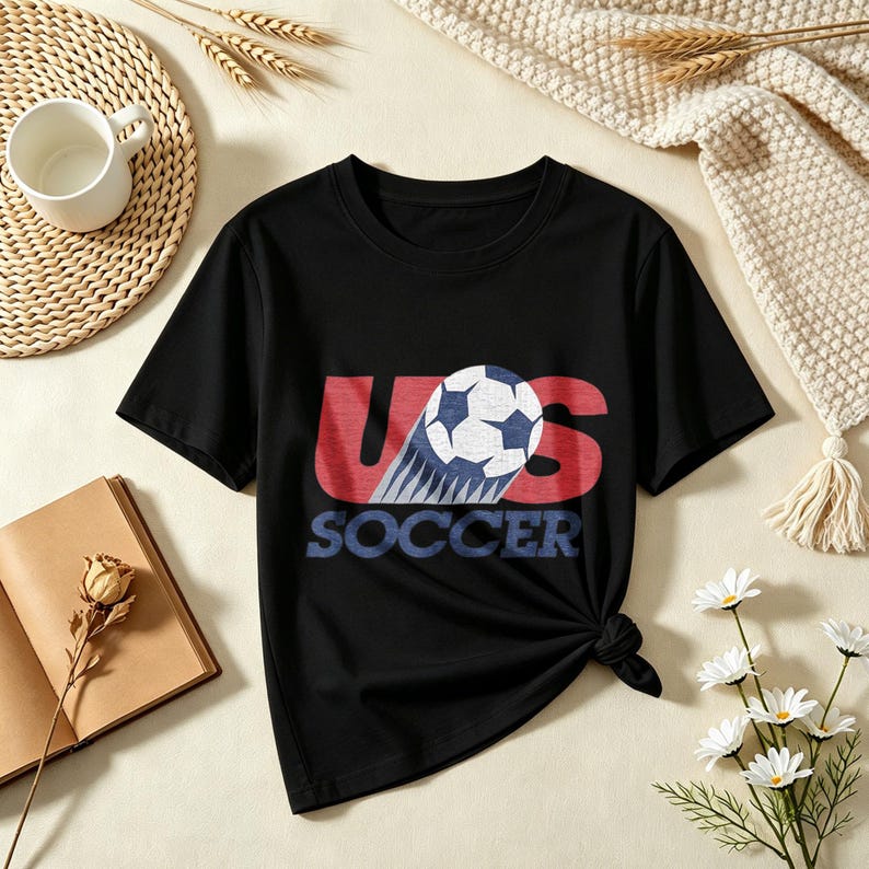 May include: A black short-sleeved t-shirt featuring a graphic print on the chest. The design displays the letters US in red with a soccer ball replacing the letter O, and the word SOCCER printed in blue underneath the graphic.