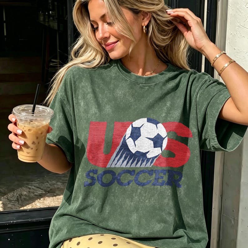 May include: A mineral-washed olive green short-sleeve t-shirt featuring a graphic print with the red text US, a black and white soccer ball, and the blue text SOCCER. The design has a distressed, vintage-inspired texture across the front.