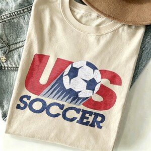 May include: A cream-colored t-shirt featuring a graphic print with the red letters U and S. A black and white soccer ball is positioned between the letters, with motion lines trailing behind it. The word SOCCER is printed in blue below the graphic.