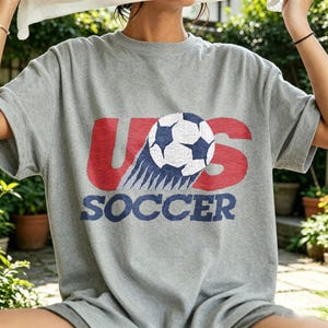 May include: A heather gray short-sleeve t-shirt featuring a graphic print with the red text US, a white and blue soccer ball illustration, and the blue text SOCCER below. The design has a distressed, vintage-inspired texture throughout the print.