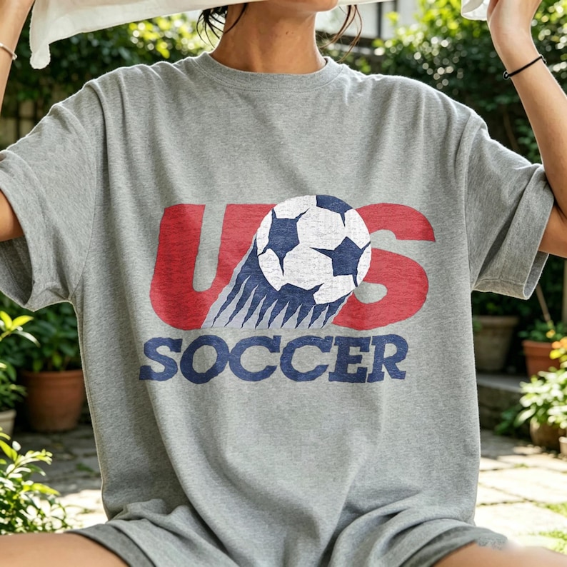 May include: A heather gray short-sleeve t-shirt featuring a graphic print with the red text US, a white and blue soccer ball illustration, and the blue text SOCCER below. The design has a distressed, vintage-inspired texture throughout the print.