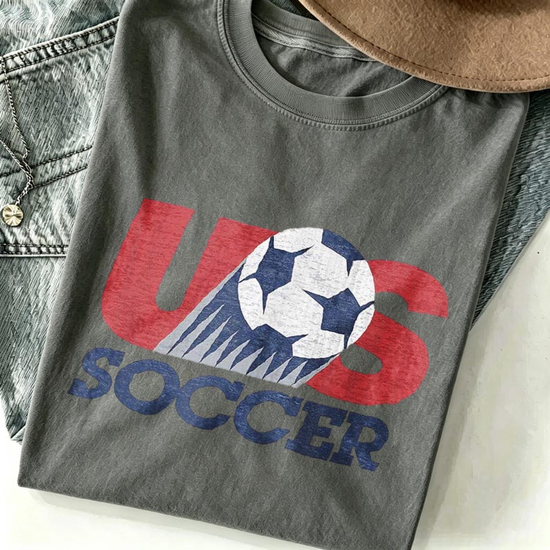 May include: A charcoal gray crew neck t-shirt featuring a distressed graphic print. The design includes the red letters US with a white and blue soccer ball replacing the letter O, and the word SOCCER printed in blue block letters below.