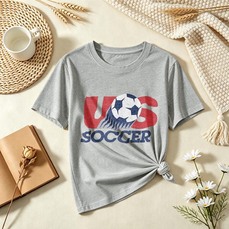 May include: A heather gray short-sleeve t-shirt featuring a graphic print of a soccer ball in motion over the red text US and the blue text SOCCER. The design has a distressed, vintage-inspired texture and is centered on the chest of the shirt.