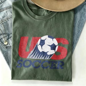May include: An olive green crew neck t-shirt featuring a distressed graphic print. The design displays the red text US with a white and blue soccer ball graphic integrated into the letters, and the word SOCCER printed in blue underneath.