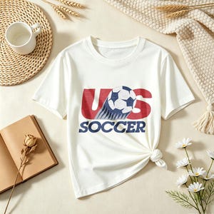 May include: A white short-sleeved t-shirt featuring a graphic print on the chest. The design displays the red text US with a soccer ball replacing the letter S, and the word SOCCER printed in blue underneath in a bold, sans-serif font.