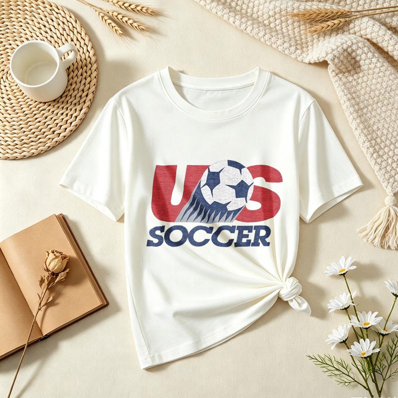 May include: A white short-sleeved t-shirt featuring a graphic print on the chest. The design displays the red text US with a soccer ball replacing the letter S, and the word SOCCER printed in blue underneath in a bold, sans-serif font.