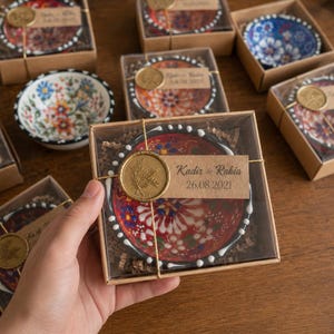 Peut inclure: Un petit bol en céramique peint à la main, orné d'un motif floral rouge, blanc et bleu, présenté dans une boîte cadeau en carton avec couvercle transparent. Une étiquette en papier kraft indique Kadir & Rabia 26.08.2021, scellée à la cire dorée.