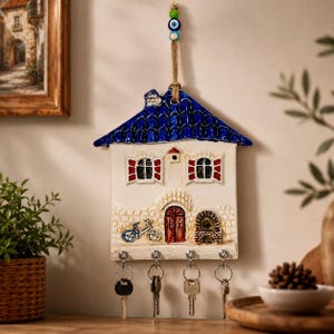 Pode incluir: Um porta-chaves de parede em cerâmica com formato de casa, telhado de telhas azuis, paredes brancas, persianas vermelhas e porta vermelha. Apresenta uma bicicleta azul e um forno de tijolos, suspenso por corda de juta com contas verdes.