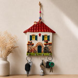 Op de afbeelding: Een keramische wand-sleutelhouder in de vorm van een huis met een rood dak, blauwe luiken en een stenen gevel. Het hangt aan een koord met een blauwe boze oog kraal en heeft vier metalen haken waaraan diverse sleutels en sleutelhangers hangen.