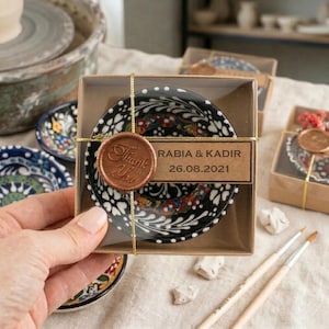 Könnte beinhalten: Eine kleine, schwarz-weiße Keramikschale mit floralem Design, präsentiert in einer transparenten Box. Die Box ist mit goldenem Faden gebunden und enthält ein braunes Etikett mit den Namen "RABIA & KADIR" und dem Datum "26.08.2021". Ein "Danke"-Siegel ist angebracht.