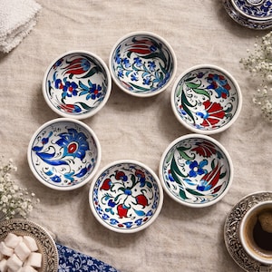 May include: A set of seven small, round ceramic bowls with white rims. Each bowl features a unique floral design in red, blue, and green on a white background. The bowls are arranged in a circular pattern on a neutral-toned surface.