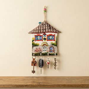 May include: A ceramic house-shaped key holder with a brown tiled roof, colourful windows, and a painted bicycle. The key holder has four hooks with keys hanging from them. A string with beads is attached for hanging.