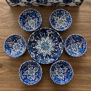 Puede incluir: Un juego de siete cuencos de cerámica azules y blancos con un diseño floral. Los cuencos están dispuestos sobre una superficie de madera, con un cuenco más grande en el centro y seis cuencos más pequeños alrededor. Los cuencos tienen un borde festoneado.
