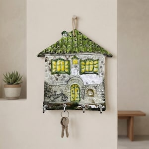 May include: A ceramic house-shaped key holder with a green tiled roof and stone-like facade. The house features yellow-lit windows and a door, with a bicycle illustration. It has four key hooks and a set of keys hanging from one. The key holder is mounted on a wall.