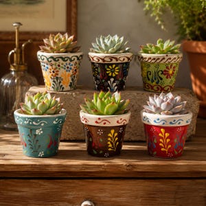 Pode incluir: Seis pequenos vasos de cerâmica pintados à mão com suculentas. Os vasos são pintados com desenhos florais e abstratos em várias cores, incluindo azul, vermelho, amarelo e verde. As suculentas têm folhas verdes e roxas. Os vasos estão dispostos sobre uma superfície de madeira.
