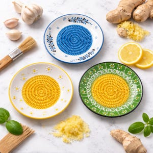 May include: Three ceramic grater plates in blue, yellow, and green, each with a textured grating surface. The plates are decorated with floral patterns and are surrounded by ingredients like garlic, ginger, and lemons, suggesting culinary use.