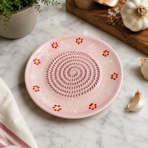 May include: A round ceramic grater plate in a soft pink hue, featuring a central spiral texture for grating and a decorative border of small red and yellow flowers with white painted leaves arranged around the perimeter of the plate.