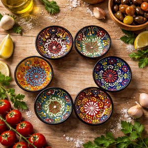 Könnte beinhalten: Ein Set aus sieben kleinen, runden Keramikschalen mit aufwendigen Blumenmustern in verschiedenen Farben, darunter Rot, Blau, Gelb und Grün. Jede Schale hat einen schwarzen Rand und ist kreisförmig angeordnet. Die Schalen stehen auf einer Holzoberfläche.