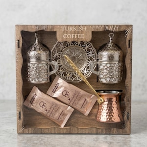 Puede incluir: Un juego de café turco en una caja de madera. El juego incluye dos tazas plateadas, un plato decorativo, una cafetera de cobre, una cuchara dorada y dos paquetes de café. La caja lleva la etiqueta "TURKISH COFFEE".