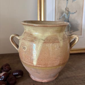 May include: A terracotta pot with two side handles, featuring a wide mouth and a tapered base. The exterior has a natural clay finish with a pale yellow glaze dripping from the rim down the sides, creating a textured and layered appearance.