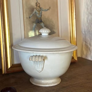 May include: A white ceramic lidded tureen with a rounded body, a pedestal base, and two decorative shell-shaped handles on the sides. The lid features a matching shell-shaped finial at the center. The surface is smooth with a glossy finish.