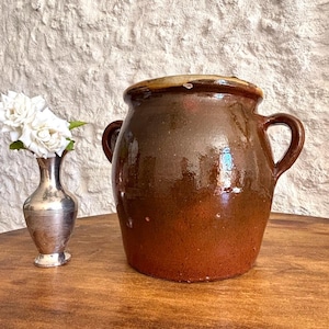 May include: A large, round, dark brown glazed ceramic crock with two side handles and a thick rim, positioned next to a small, slender, metallic silver-toned vase containing two white flowers with visible petals and green leaves.