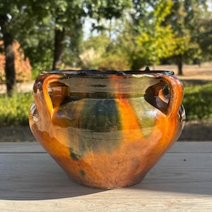 May include: A glazed ceramic pot featuring a rounded body, a wide opening with a scalloped rim, and two side handles. The exterior surface displays a mottled colour palette of deep amber, burnt orange, and dark forest green in a glossy finish.