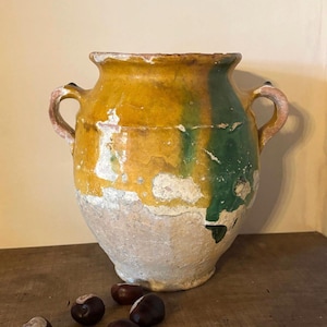 May include: A rustic earthenware vessel featuring two side handles, a wide mouth, and a bulbous body. The upper portion is finished with a yellow and green glazed surface showing significant wear, while the lower base remains unglazed and textured.