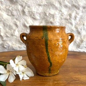 May include: A small, round, ochre-colored ceramic pot with two side handles and a single vertical green stripe running down the center. The surface has a glossy, uneven glaze with visible texture and a wide opening at the top.