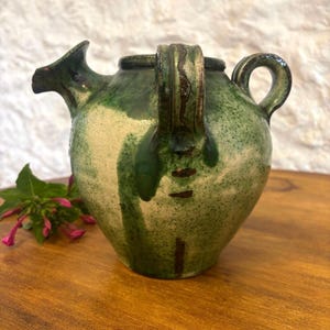 May include: A round ceramic pitcher with a mottled green and cream glaze. The vessel features a short spout, a thick vertical handle with dark brown linear accents, and a smaller side handle. The surface has a textured, rustic appearance.