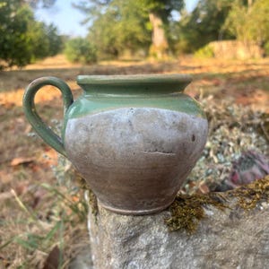 May include: A small, round ceramic pot with a single curved handle. The upper portion features a smooth, glossy green glaze, while the lower body has a matte, textured, light beige finish with visible surface imperfections and a tapered base.