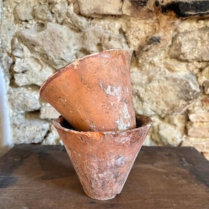 Puede incluir: Dos macetas de arcilla de color terracota desgastadas, con forma cónica. Las macetas están apiladas, revelando su textura envejecida y las variaciones naturales de la arcilla. El fondo presenta un muro de piedra rústico.