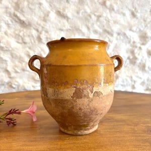 May include: A small, two-handled earthenware pot featuring a glossy mustard-yellow glaze on the upper portion and a textured, unglazed terracotta base. The vessel has a wide rim and a rounded body, showing signs of wear and surface weathering.