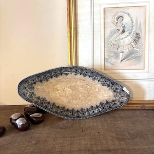 May include: An oval ceramic dish featuring a dark blue intricate floral and scrollwork border pattern surrounding a textured, cream-colored center. The dish has a slightly weathered appearance with a small chip visible on the right edge.