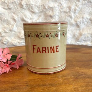 Puede incluir: Un recipiente cilíndrico de cerámica con un acabado de esmalte craquelado de color crema. La palabra FARINE está impresa en letras rojas en la parte frontal, acentuada por una banda horizontal de pequeñas flores rojas y enredaderas verdes.