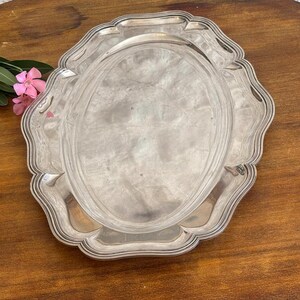 May include: An oval silver-toned serving tray with a polished, reflective surface and a decorative scalloped edge featuring multiple concentric ridges. The tray has a smooth, flat central area and a wide, ornate border with a wavy, curved rim.