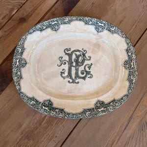 Könnte beinhalten: Eine ovale Servierplatte aus Keramik mit cremefarbener Basis und einem dunkelgrünen Zierrand mit Blumen- und Tiermotiven. In der Mitte der Platte befindet sich ein verziertes, dunkelgrünes, verschlungenes Monogramm der Buchstaben P und L.