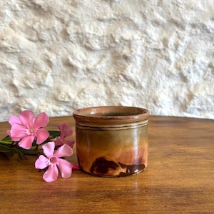 May include: A small, cylindrical ceramic pot with a glazed finish in shades of brown, tan, and dark amber, featuring a distinct rim. Beside the pot are two vibrant pink oleander flowers with five petals each, displayed on a wooden surface.